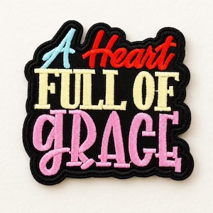Puede incluir: Un parche bordado negro con el texto A Heart FULL OF GRACE. La palabra A es azul claro, Heart es roja, FULL OF es amarilla y GRACE es rosa. El parche tiene un borde negro festoneado y un acabado de tela texturizada.