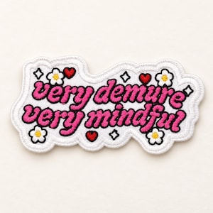 Può includere: Una toppa ricamata con sfondo bianco e bordo smerlato. Il testo "very demure very mindful" è scritto in lettere rosa in grassetto. Il design è decorato con piccoli fiori bianchi con centro giallo, cuori rossi e contorni di stelle nere.