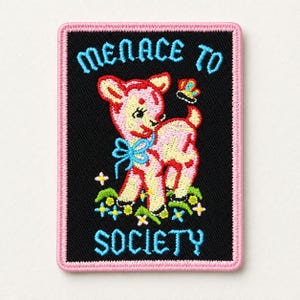 Könnte beinhalten: Ein rechteckiger bestickter Aufnäher mit schwarzem Hintergrund und rosa Rand. Er zeigt ein rosa Rehkitz mit blauer Schleife, einen kleinen Schmetterling und bunte Blumen. Der Text lautet "MENACE TO SOCIETY" in hellblauen Stickbuchstaben.