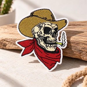 Puede incluir: Un parche bordado que muestra una calavera con un sombrero de vaquero color canela y un pañuelo rojo alrededor del cuello. La calavera tiene un cigarrillo amarillo en la boca con un fino rastro de humo blanco, todo bordeado en hilo blanco.
