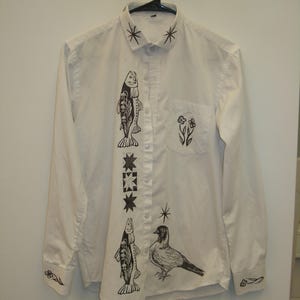 May include: A white long-sleeve button-down shirt featuring black ink-style illustrations. The design includes two fish, a bird, star shapes on the collar, and floral motifs on the chest pocket and cuffs, all printed on a crisp white fabric base.