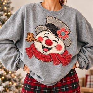 Könnte beinhalten: Ein graumeliertes Sweatshirt mit Rundhalsausschnitt und einer großen Applikation aus ausgefranstem Stoff, die einen lächelnden Schneemann zeigt. Der Schneemann trägt einen dunkelgrauen Zylinder mit roter Blume, einen roten Schal und eine braune Pfeife.