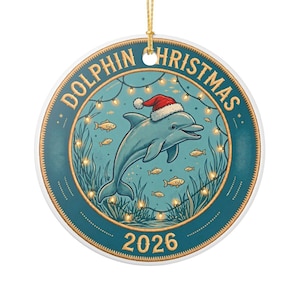 May include: A round ceramic ornament featuring a blue dolphin wearing a red and white Santa hat, surrounded by small fish, seaweed, and string lights. The text reads DOLPHIN CHRISTMAS and 2026. It includes a gold hanging string at the top.