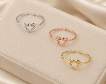 May include: Three open-style rings featuring a heart-shaped knot design. The set includes one silver-toned ring, one rose gold-toned ring, and one gold-toned ring. Each ring has a thin, smooth band with a small spherical detail at the open ends.