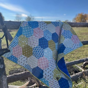 May include: A patchwork quilt with a hexagonal design, displayed on a wooden fence. The quilt showcases blue, pink, and yellow geometric shapes with diverse patterns. It has a yellow border and a solid blue reverse side. The quilt is a handmade item.