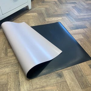 May include: A black rectangular mat with a light gray top layer, partially rolled back to reveal the black underside. The mat is on a wooden floor with a herringbone pattern. The background includes a white cabinet.