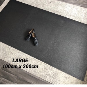 Puede incluir: Una alfombra rectangular negra grande, de 100cm x 200cm, con una superficie texturizada. Un par de zapatos de cuero negro descansa sobre la alfombra. La palabra "LARGE" está impresa en la alfombra.
