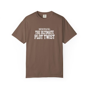 May include: A brown short-sleeve t-shirt featuring the white text "MENOPAUSE: THE ULTIMATE PLOT TWIST" printed in a bold, block-style font across the center of the chest. The garment has a crew neckline and a standard fit.