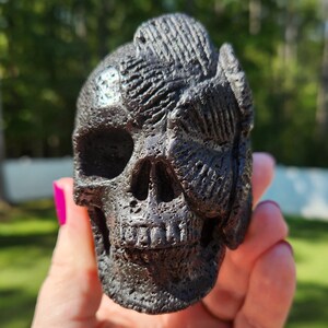 May include: A dark, textured skull sculpture with detailed features. The skull is held in a hand, with a blurred green background. The sculpture has a rough, carved appearance, with visible eye sockets, nasal cavity, and teeth.