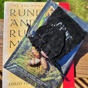 Op de afbeelding: Een set runestones in een zwarte fluwelen zakje, met een boek getiteld "The Big Book of Runes and Rune Magic" op de achtergrond. De boekomslag toont een boom met een groene achtergrond.