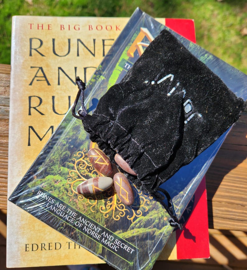 Op de afbeelding: Een set runestones in een zwarte fluwelen zakje, met een boek getiteld "The Big Book of Runes and Rune Magic" op de achtergrond. De boekomslag toont een boom met een groene achtergrond.