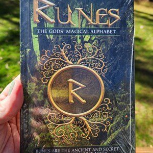 Op de afbeelding: Een boek getiteld "Runes: The Gods' Magical Alphabet" met een groene en gouden omslag met een runesymbool in een cirkel omgeven door een rankontwerp. De tekst "Runes are the ancient and secret language of Norse magic" staat gedrukt op de onderkant van de omslag.