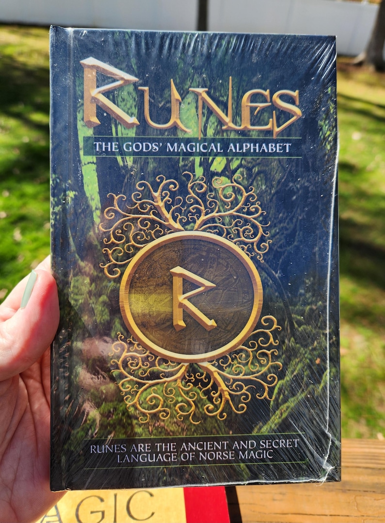 Op de afbeelding: Een boek getiteld "Runes: The Gods' Magical Alphabet" met een groene en gouden omslag met een runesymbool in een cirkel omgeven door een rankontwerp. De tekst "Runes are the ancient and secret language of Norse magic" staat gedrukt op de onderkant van de omslag.