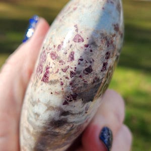 Könnte beinhalten: Ein polierter, oval geformter Edelstein mit einer Mischung aus Farben. Der Stein hat eine Basis aus hellgrau und beige, mit weißen Streifen und Flecken aus tiefroten Kristallen. Die Oberfläche ist glatt und reflektierend, gehalten in einer Hand.