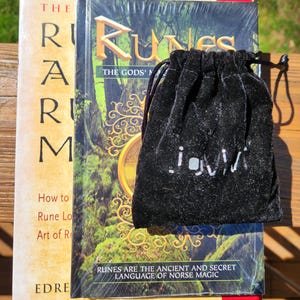 Op de afbeelding: Een zwart fluwelen zakje met een wit logo, liggend op een boek getiteld "Runes: The Gods' Magic". De boekomslag heeft een groene en gouden illustratie van een boom met een cirkelvormig ontwerp.