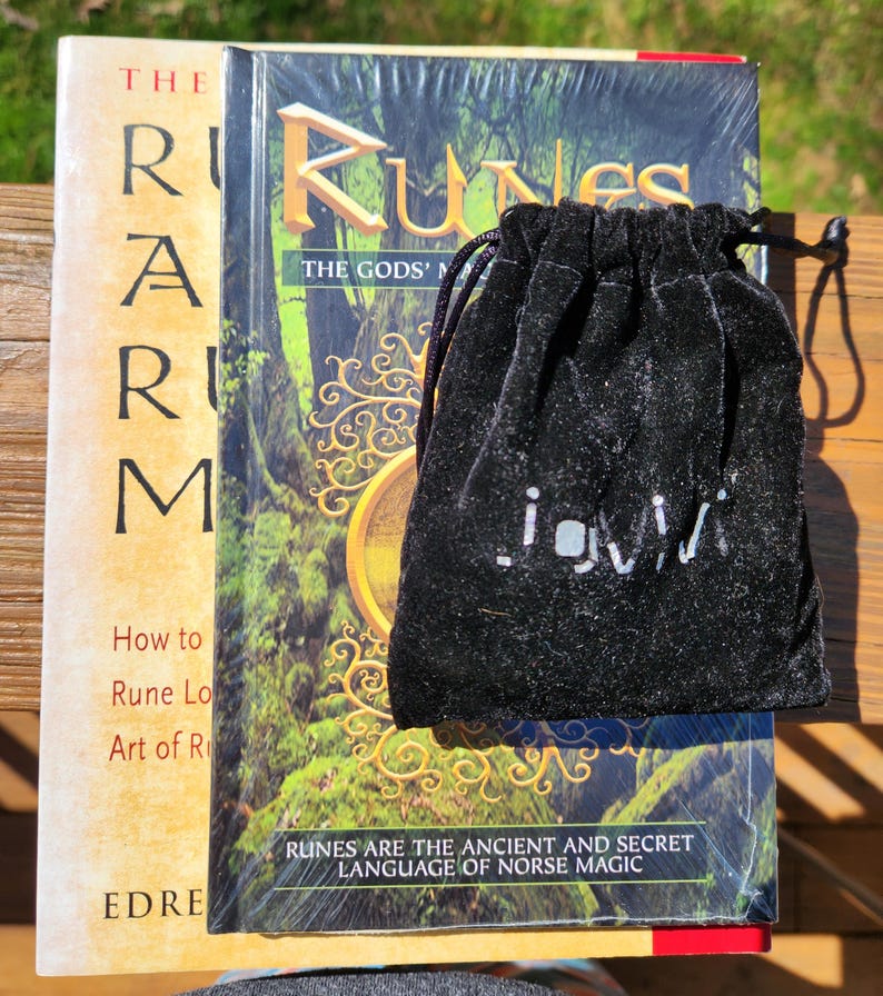 Op de afbeelding: Een zwart fluwelen zakje met een wit logo, liggend op een boek getiteld "Runes: The Gods' Magic". De boekomslag heeft een groene en gouden illustratie van een boom met een cirkelvormig ontwerp.