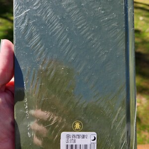 Op de afbeelding: Een groen hardback boek met een sticker op de achterkant. De sticker heeft een barcode, de tekst "ISBN: 978-07387-5381-2", "US $17.95" en een websiteadres. Het boek is verpakt in plastic.
