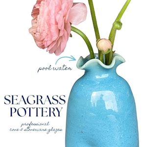 May include: A light blue stoneware vase with a ruffled rim, containing pink flowers. Text on the image reads: SEAGRASS POTTERY, pool water, and professional cone 5 stoneware glazes. The vase has a smooth, glossy finish and a rounded shape.