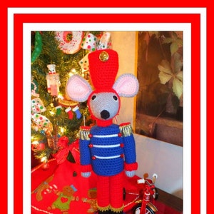 May include: A crocheted mouse dressed as a toy soldier, wearing a red hat, blue and white uniform, and red trousers. The mouse is standing on a gold and blue base, in front of a Christmas tree and a red toy tricycle.
