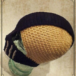 Puede incluir: Un gorro de punto de crochet con un borde negro y un cuerpo de color beige. El gorro tiene un dise&ntilde;o &uacute;nico con una m&aacute;scara facial negra y gris unida a la parte delantera.