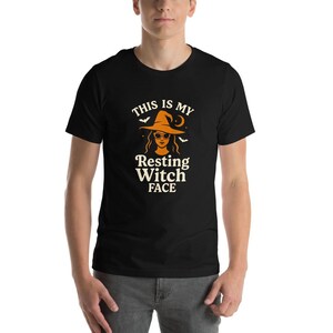 May include: A black short-sleeve t-shirt featuring a graphic of a person wearing a witch hat and sunglasses. The text reads THIS IS MY Resting Witch FACE in cream-colored lettering, accompanied by small orange bats and a crescent moon.