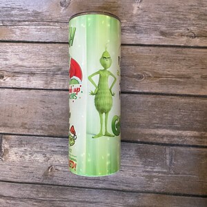 May include: A green and white tumbler featuring the Grinch character. The tumbler has text and images of the Grinch in a Santa hat and the words "Pick up Grinches" and "I am a Grinch". The tumbler is cylindrical.