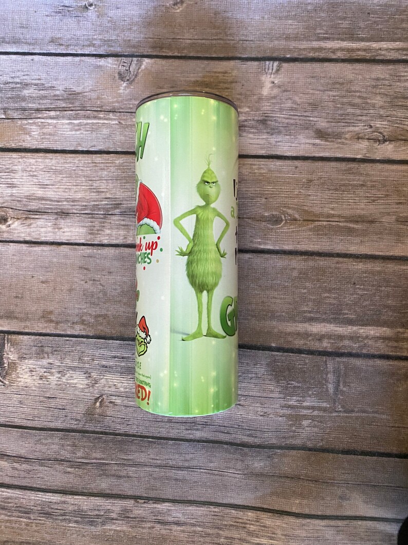 May include: A green and white tumbler featuring the Grinch character. The tumbler has text and images of the Grinch in a Santa hat and the words "Pick up Grinches" and "I am a Grinch". The tumbler is cylindrical.