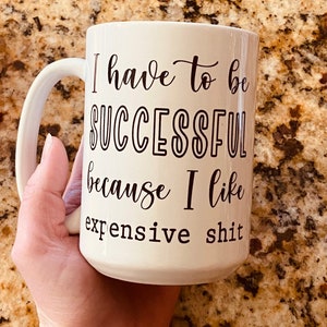 May include: White ceramic mug with a large handle, featuring the text "I have to be SUCCESSFUL because I like expensive shit" in black script. The mug is held by a hand.