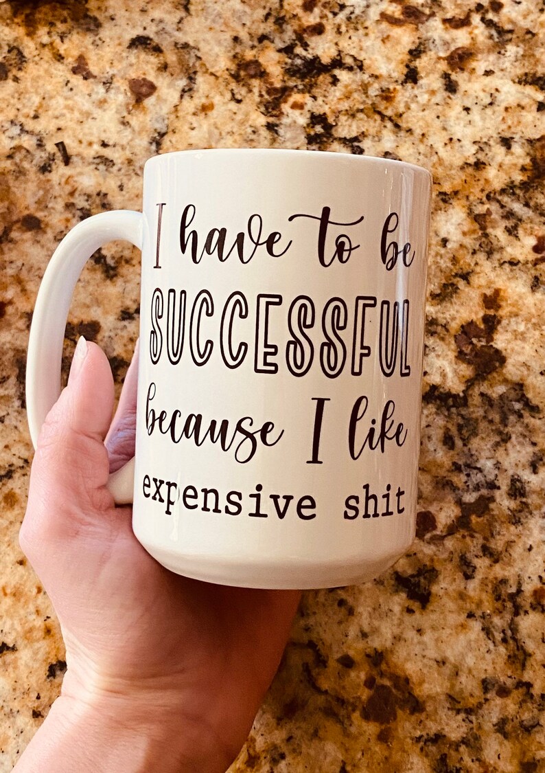 May include: White ceramic mug with a large handle, featuring the text "I have to be SUCCESSFUL because I like expensive shit" in black script. The mug is held by a hand.