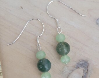 May include: A pair of dangle earrings featuring silver hooks and a combination of green beads. The earrings have a mix of round and faceted beads in varying shades of green, creating a layered effect. The earrings are displayed on a light wooden surface.