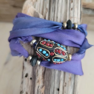 May include: A purple silk ribbon tied around a piece of driftwood with a black and gold bead with red and blue accents.