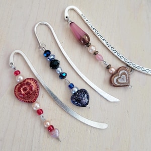May include: Three silver bookmark pendants with decorative charms. The charms include a pink glass teardrop, a blue glass heart, and a red heart with a floral design. Each charm is attached to a silver chain with beads.