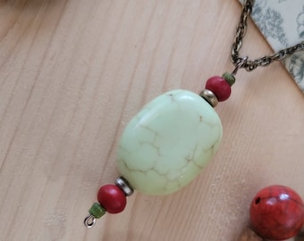 Op de afbeelding: Een ketting met een groene stenen hanger en rode en groene kralen. De ketting ligt op een houten oppervlak met andere kralen.