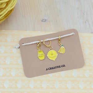 Op de afbeelding: Een set van drie gele emaille bedels op goudkleurige ringen, met een melkpak, een citroen en een banaan. De bedels zijn bevestigd aan een bruine kaart met de tekst "A CREATIVE CO."