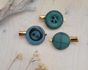 May include: Three green and blue button hair clips with gold metal clasps. The buttons are different designs, one is a solid green with a woven pattern, one is a solid green with four holes, and one is a blue button with a black swirl design.