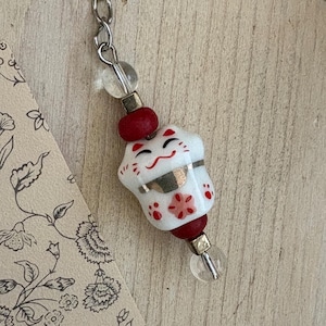 May include: A white ceramic Maneki Neko charm with red detailing and a smiling face, strung on a silver chain with clear and red beads. The charm features a floral design and is a symbol of good luck.