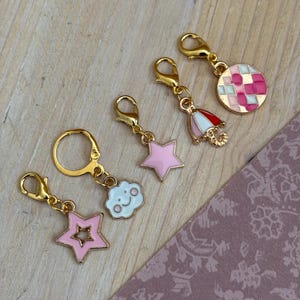 Könnte beinhalten: Eine Sammlung goldfarbener Anhänger mit verschiedenen Designs, darunter ein rosa Stern, eine lächelnde Wolke, ein rot-weißer Regenschirm und ein rosa-weiß karierter Kreis. Die Anhänger liegen auf einer hellen Holzoberfläche.