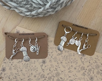 May include: Silver-toned knitting stitch markers with charms shaped like yarn, scissors, and buttons are displayed on brown cards. A large ball of gray and white yarn sits above the cards. The cards are labeled "A CREATIVE CO."