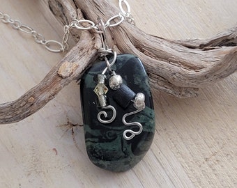 Op de afbeelding: Een zilveren ketting met een hanger van groene en zwarte steen. De hanger heeft zilveren draadaccenten en zwarte kralen.
