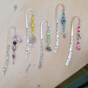 Puede incluir: Cinco colgantes de marcapáginas de plata con cuentas y abalorios de diferentes colores. Cada marcapáginas presenta un diseño floral y un pequeño abalorio en la parte inferior.