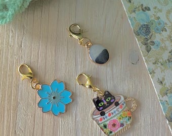 Puede incluir: Tres colgantes dorados con cierres de mosquetón. Uno es una flor azul claro, otro un óvalo blanco y negro, y el tercero un gato negro en una taza de té floral. Los colgantes están sobre una superficie de madera clara.