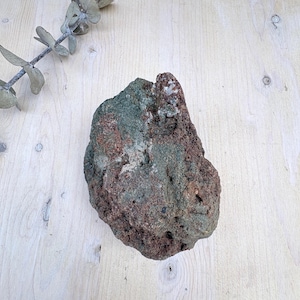 May include: A rough, irregularly shaped rock featuring a mottled surface with deep forest green and earthy reddish-brown tones. The stone has a porous, uneven texture with visible mineral deposits and a jagged, natural formation throughout its body.