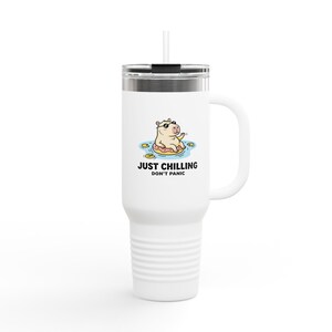 Op de afbeelding: Een witte roestvrijstalen beker met handvat, deksel en een doorzichtig rietje. De voorzijde toont een afbeelding van een capibara met zonnebril op een roze donut-zwemband in blauw water, met de tekst "JUST CHILLING DON'T PANIC" eronder.