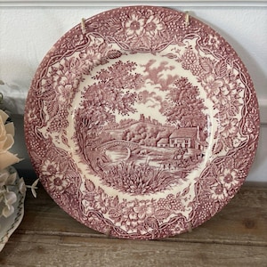 May include: A round ceramic plate featuring a maroon and cream transferware pattern. The center depicts a rural landscape with a stone bridge, a house, and a person on horseback. The wide border is decorated with detailed floral and berry motifs.