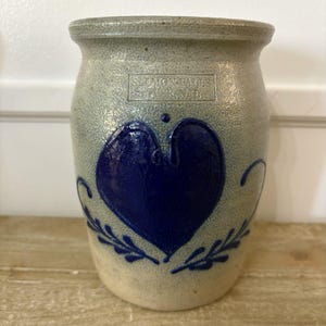 May include: A gray stoneware crock featuring a deep blue painted heart design on the front. The vessel has a flared rim and includes the stamped text SALMON FALLS DOVER, N.H. above the heart, with blue leaf accents below the heart.