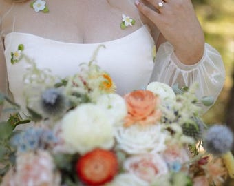 May include: A white dress featuring a sheer illusion neckline and long sleeves adorned with small, embroidered white flower appliqués with green leaves. The bodice has a straight-across neckline, and the garment is paired with a large floral bouquet.