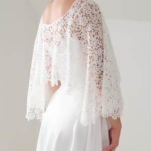 Könnte beinhalten: Ein weißes Spitzen-Capelet mit floralem Muster. Das Capelet hat einen runden Ausschnitt und fällt über die Schultern. Das Kleidungsstück besteht aus zarter, offen gewebter Spitze und wird über einem weißen Kleid getragen.