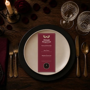 May include: A rectangular menu card titled Midnight Masquerade with a gold mask icon and clock graphic, placed on a white plate atop a black charger. The menu lists Burrata with Roasted Figs, Filet au Poivre, and Midnight Chocolate Torte.