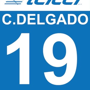 Può includere: Un cartello rettangolare blu con il testo bianco C.DELGADO sopra il grande numero 19 in bianco. In alto, una barra orizzontale bianca contiene un logo blu con tre linee e il testo TELCEL in un carattere blu in grassetto.