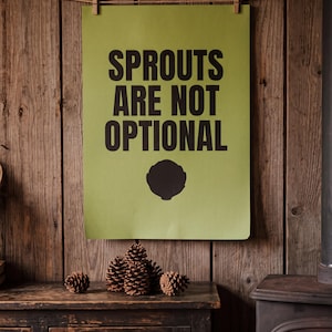 May include: A rectangular olive green poster featuring the black text SPROUTS ARE NOT OPTIONAL in a bold sans-serif font, positioned above a black silhouette of a single sprout. The poster is suspended by two wooden clips on a string.